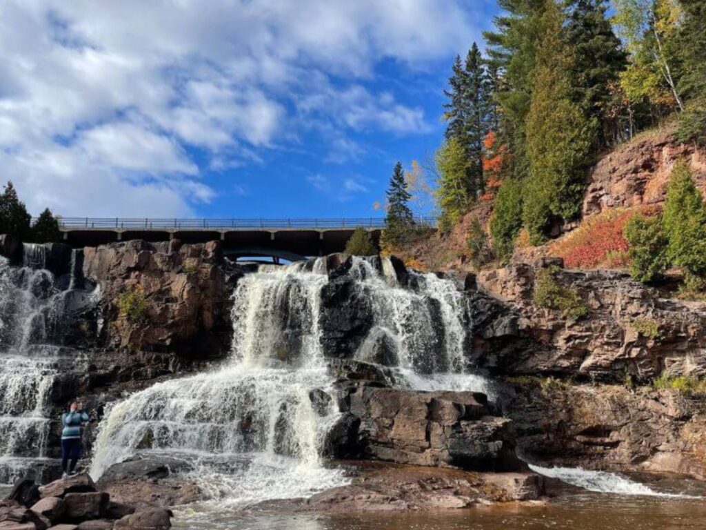 Gooseberry Falls dropping down a rocky cliff underneath a bridge on a day with blue skies.